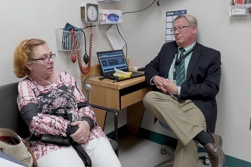 Denise Brown, who suffers from aortic stenosis and needed heart valve replacement surgery, sits with Dr. John Conte at Penn State Milton S. Hershey Medical Center. Brown is sitting in an exam room on a chair next to Conte, who is sitting on a stool next to a desk on which his laptop sits, open. Brown wears a cannula in her nose to assist with breathing.