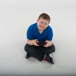 A young boy sits on the floor holding a video game controller with both hands, looking up at the camera, smiling.