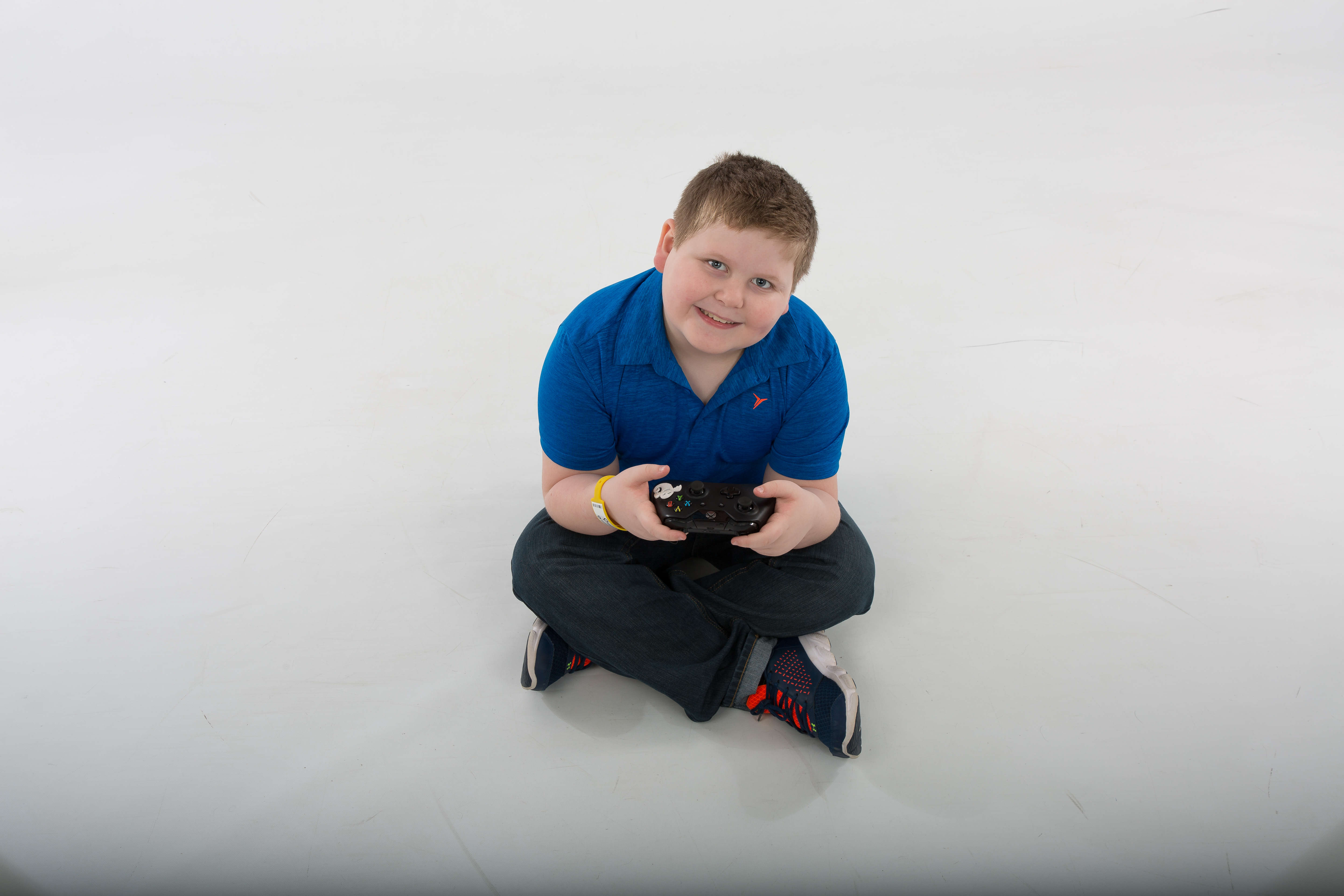 A young boy sits on the floor holding a video game controller with both hands, looking up at the camera, smiling.