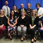 Peter Carlino, back row, center, poses with the 2018 winners of the Elizabeth Powers Carlino Nursing Excellence Scholarship Award. Three women and one man stand with Carlino in the back row. Five women sit on chairs in the front row. They are dressed in business casual attire. Behind them is a blue curtain.