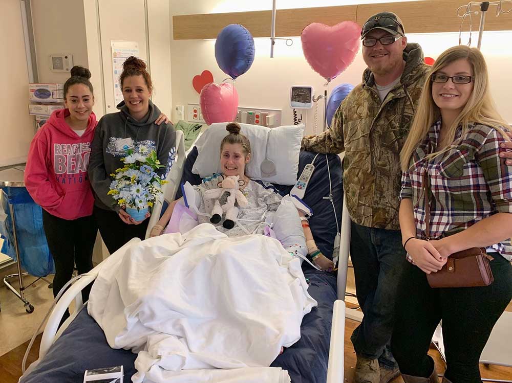 Dina Anthony, center, a patient at Hershey Medical Center, cries tears of joy during her daughter Brittnay’s gender reveal party. She is propped up in a hospital bed, is wearing a hospital gown and has a stuffed animal on her chest. Brittnay, second from left, holds flowers that show she will have a baby boy. She is wearing a gray hoodie sweatshirt. On the far left is another young woman wearing a hoodie sweatshirt. Second from right is a man wearing a camouflage jacket. On the far right is a woman with long, blonde hair and a plaid shirt. Behind them are balloons.