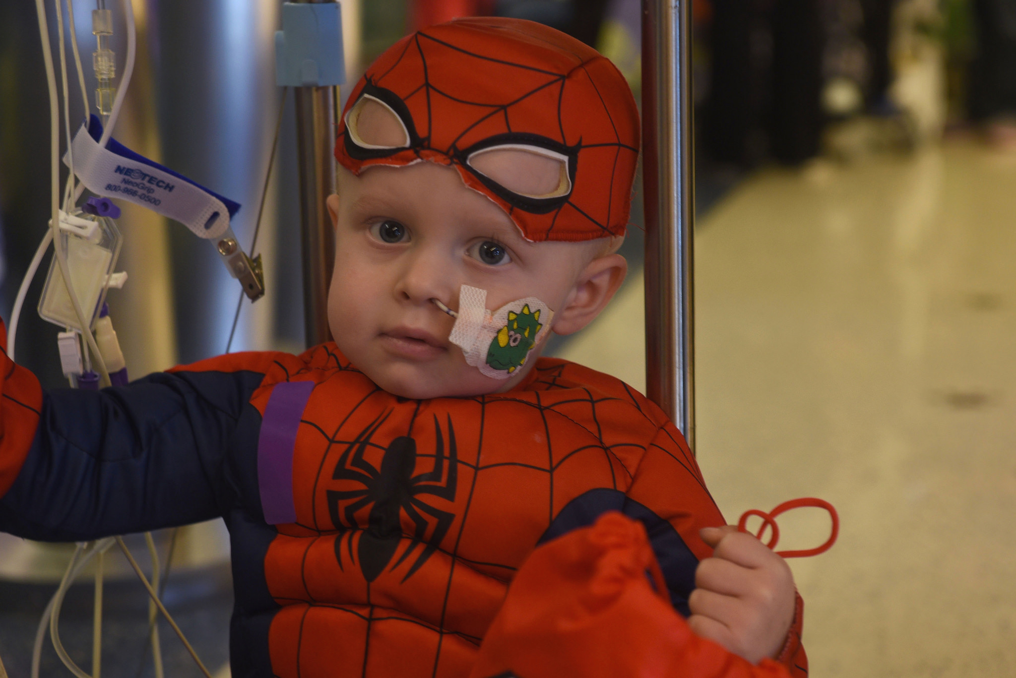 A young child in a Spiderman costume poses for a photo while sitting on the base of an IV pole. Some IV cords are in the background. The child holds a red bag with one hand.