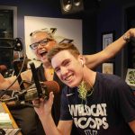 A teenage boy sits at a microphone in a radio studio, smiling. A woman stands behind him with arms outstretched, also smiling.