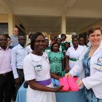 Penn State College of Medicine Global Health Scholars student Becky Koob presents a pair of pink hospital-grade shoes to Gladys Ampomah-Ababio, head of nursing at Eastern Regional Hospital, Ghana, outside the hospital building. Koob wears a white lab coat over her dress, and Ampomah-Ababio wears a white nurse’s uniform with a purple belt. Other hospital staff stand behind a table of donated shoes, smiling.