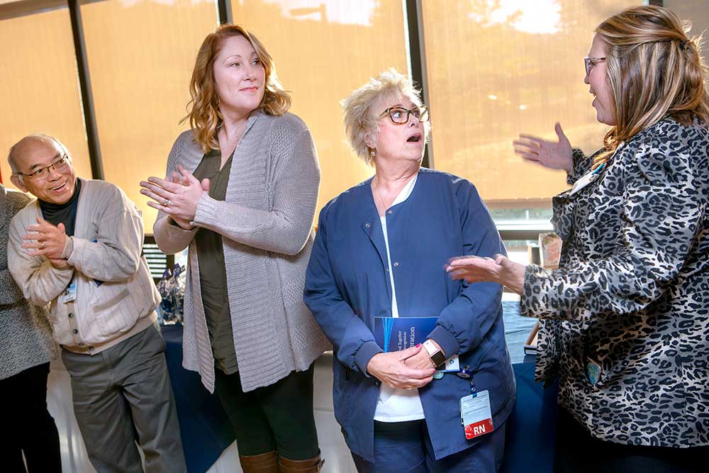 Barbara Brehm of Employee Health, second from right, looks shocked after being named Employee of the Year. She is wearing a blue jacket and pants. From left, Employees of the Month Alan Lam, wearing a button-down sweater and slacks, and Ellie Hogentogler, wearing a long sweater, top, pants and boots, clap and Amanda Hulse, wearing a leopard jacket and pants, puts her arms out toward Brehem. Behind them are windows covered with shades.