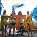 Four teenage girls hold oversized letters that spell out "FTK." Blue and white balloons and a window are in the background.