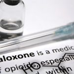 A small bottle and covered syringe needle are placed on a piece of paper with typed words that include “Naloxone, “medication” and “opioids.”