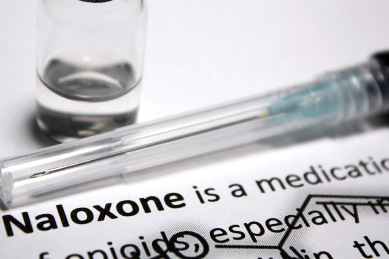 A small bottle and covered syringe needle are placed on a piece of paper with typed words that include “Naloxone, “medication” and “opioids.”