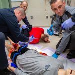 Three Pennsylvania State Police cadets learn how to apply a tourniquet during Stop the Bleed training. A man in the front-left tightens a tourniquet strap on a dummy, which has a fake gunshot wound on its right leg and blood on its left leg. A man in the back-left holds a cloth to the back of an actor. A man in the front-left reaches for a device with his gloved hand, which is covered in fake blood. The arm of another cadet reaches in holding a device. They are working in a clinical room with a vinyl floor and a curtain on the left.