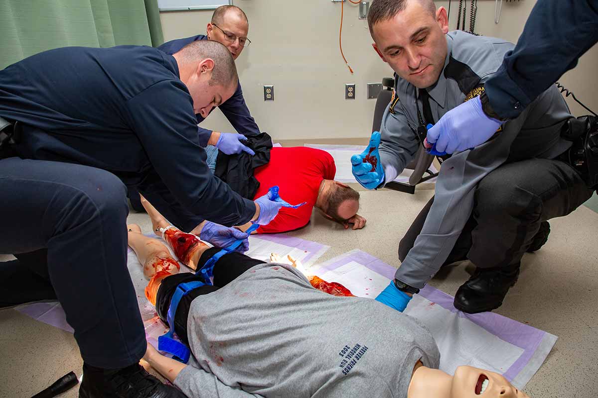 Three Pennsylvania State Police cadets learn how to apply a tourniquet during Stop the Bleed training. A man in the front-left tightens a tourniquet strap on a dummy, which has a fake gunshot wound on its right leg and blood on its left leg. A man in the back-left holds a cloth to the back of an actor. A man in the front-left reaches for a device with his gloved hand, which is covered in fake blood. The arm of another cadet reaches in holding a device. They are working in a clinical room with a vinyl floor and a curtain on the left.