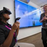 Deajanee Lee, a Milton Hershey School student, looks through virtual reality goggles during the Penn State Health Milton S. Hershey Medical Center STEM Goes Red event. Eliza Donne, a multimedia specialist at the Harrell Library, watches a screen. Behind them is a screen with video of stingrays swimming in the ocean. Deajanee is wearing a black top and braids. Eliza is wearing glasses and a black and white top.