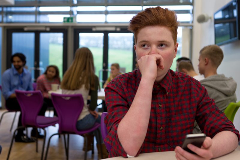 A teenage boy sits at a table in a school cafeteria. Other children are seated at other tables in the background.