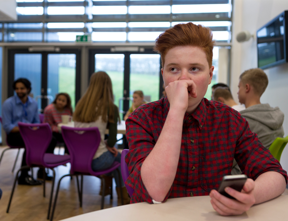 A teenage boy sits at a table in a school cafeteria. Other children are seated at other tables in the background.