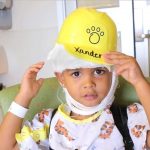Xander, a three-year-old patient at Penn State Health Children's Hospital, wears a yellow hard hat with his name and a paw print on it. He has a bandage wrapped around his head and below his chin and is wearing a hospital smock with cats on it, plus a hospital identification wristband. He is sitting in a chair. Behind him are a sink and a door.