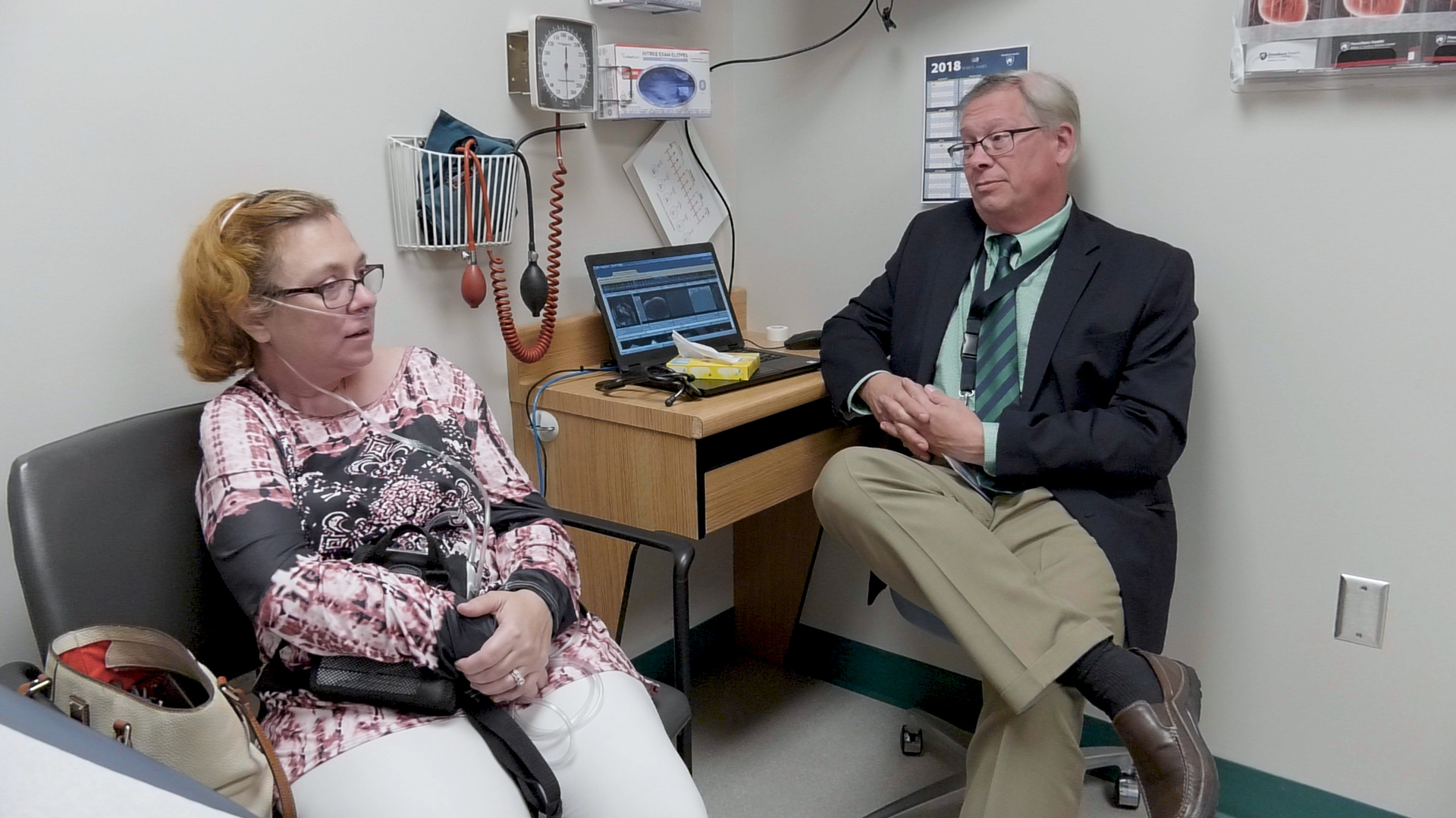 Denise Brown, who suffers from aortic stenosis and needed heart valve replacement surgery, sits with Dr. John Conte at Penn State Milton S. Hershey Medical Center. Brown is sitting in an exam room on a chair next to Conte, who is sitting on a stool next to a desk on which his laptop sits, open. Brown wears a cannula in her nose to assist with breathing.