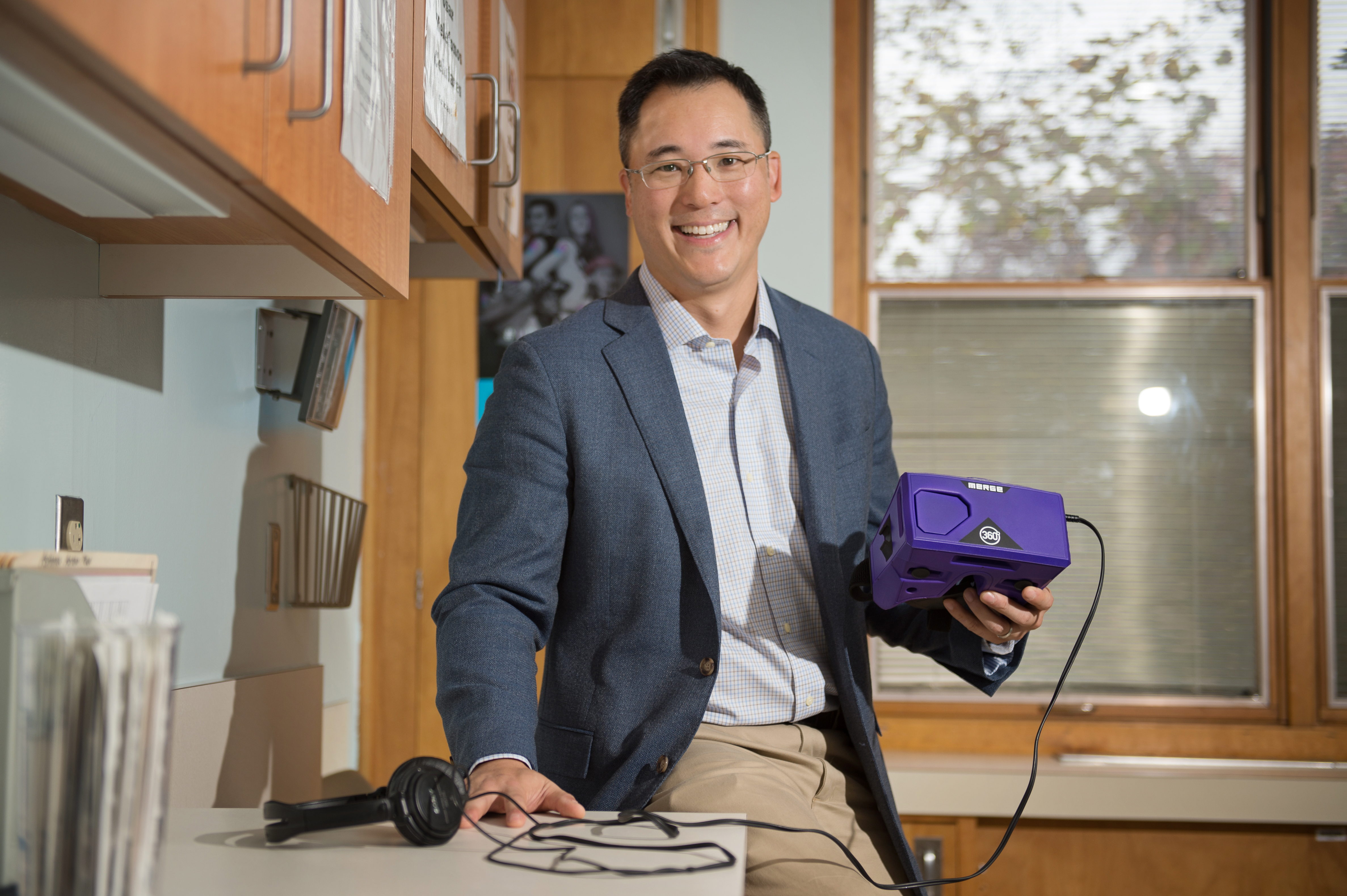 Dr. Jerry Lee, a pediatrician at Penn State Health St. Joseph Downtown Pediatric Practice, holds virtual reality goggles. The device in his left hand looks like a purple box. A cable connects it to headphones that are set on the counter. Dr. Lee is smiling and wearing glasses, a sports jacket, dress shirt and khaki pants. Behind him is a window with a shade. Next to him are wooden cabinets covered in signs. On the counter is a file stand with files.