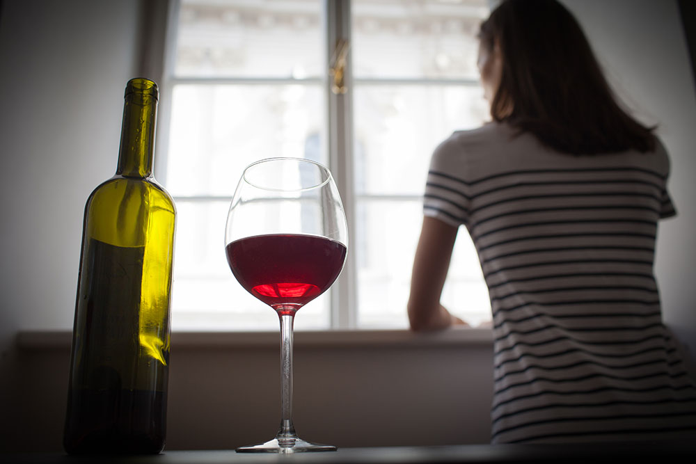A woman stares out a window. Behind her, in the foreground, a glass of wine sits next to a bottle.