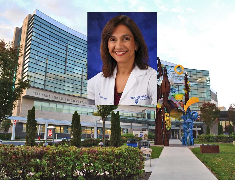 Dr. Debra Byler smiles in a head-and-shoulders professional portrait. She is wearing a white lab coat with the Penn State Health Children's Hospital logo on the right and her name on the left. She has shoulder-length hair and is wearing a sweater top and a watch.