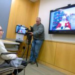 A girl with glasses smiles toward a wall-mounted TV screen showing Santa Claus while a man points a camera at her.