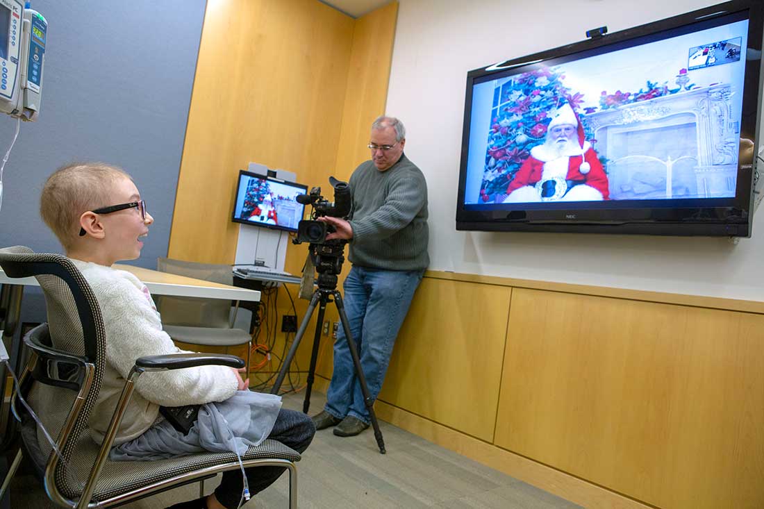 A girl with glasses smiles toward a wall-mounted TV screen showing Santa Claus while a man points a camera at her.