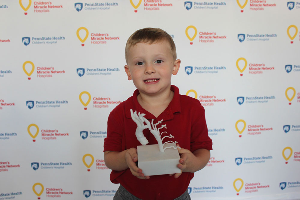 Henry Thomas, age 3, of Boiling Springs holds a 3D printed model of his own heart. He stands in front of a background with Penn State Health Children’s Hospital and Children’s Miracle Network Hospitals logos. He is wearing a polo shirt and pants. The model of his heart is on a white plastic block and resembles a gnarled tree.