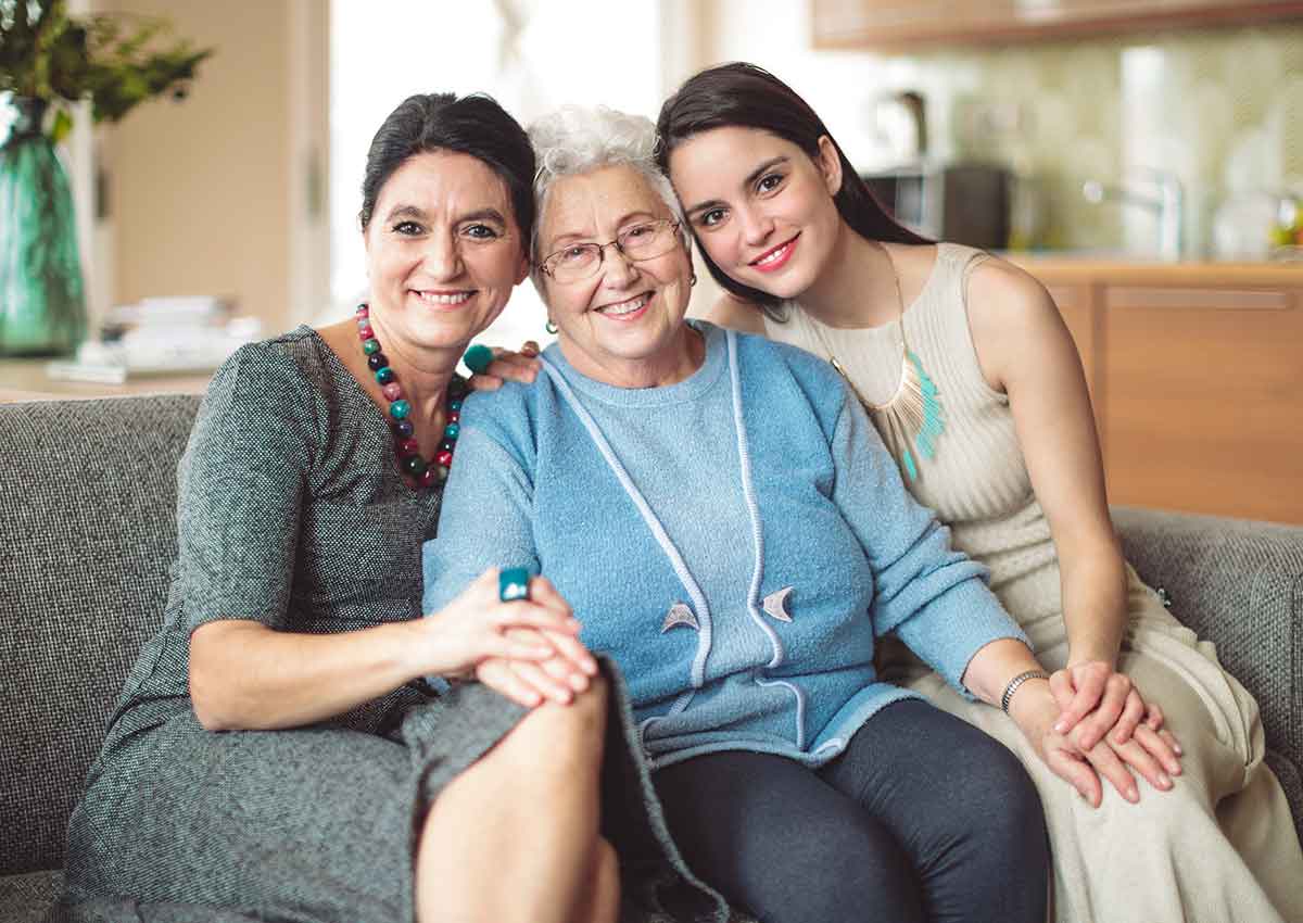 Different generations of women in a family hold hands together on a couch. In the background a vase of flowers rises from a kitchen.