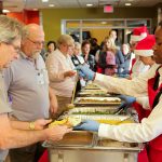 Penn State Health employees stand in a line at a buffet in the Rotunda Café during the holiday celebration. Three food service workers serve food on the right. Two are wearing Santa caps. Trays of food include turkey, mashed potatoes and green beans.