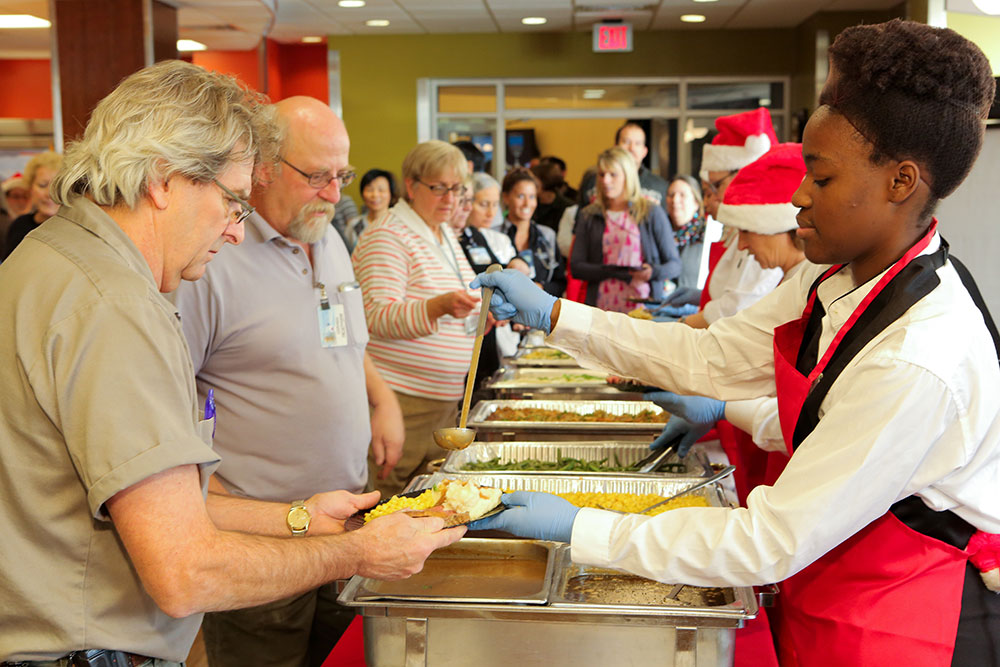 Penn State Health employees stand in a line at a buffet in the Rotunda Café during the holiday celebration. Three food service workers serve food on the right. Two are wearing Santa caps. Trays of food include turkey, mashed potatoes and green beans.