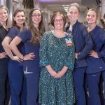 Nine women employees of the Heart and Vascular Progressive Care Unit at Hershey Medical Center pose in the middle of the unit’s hallway. Julie Werner, nurse manager, stands in the middle of the group wearing a dress and ID badge. The other women stand on either side of her angled to the side with their hands on their hips smiling.