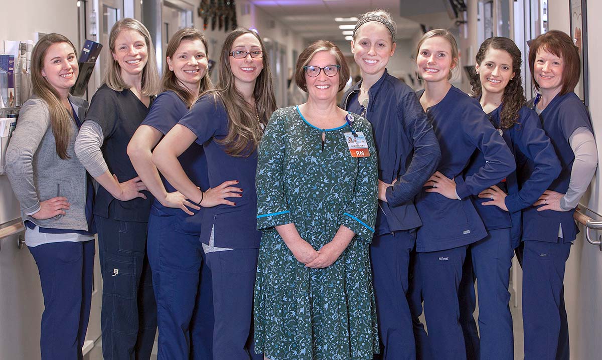 Nine women employees of the Heart and Vascular Progressive Care Unit at Hershey Medical Center pose in the middle of the unit’s hallway. Julie Werner, nurse manager, stands in the middle of the group wearing a dress and ID badge. The other women stand on either side of her angled to the side with their hands on their hips smiling.