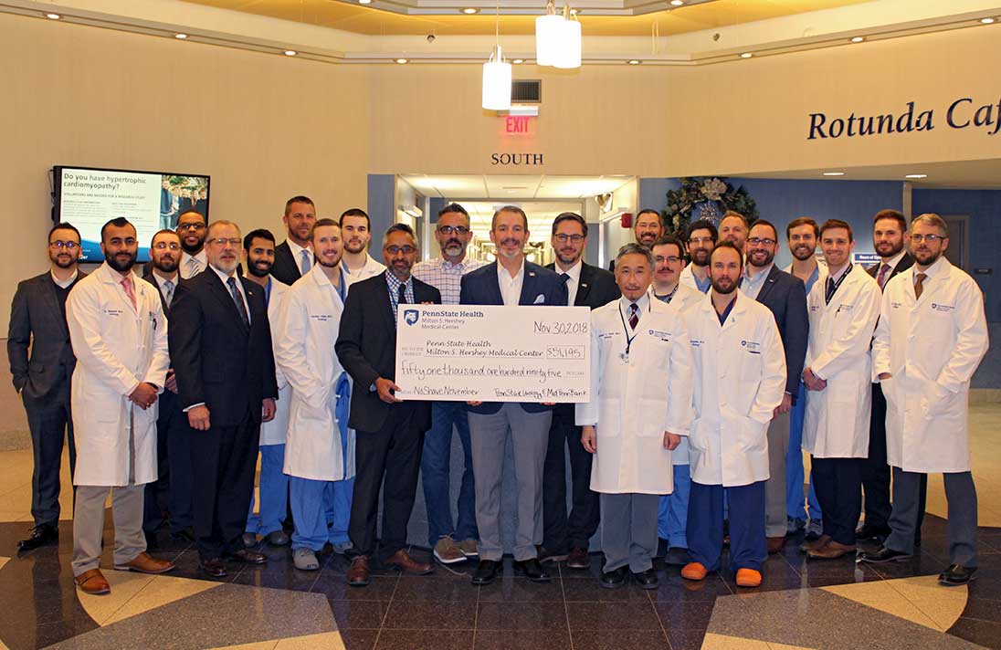 Dr. Jay Raman of the Division of Urology poses with Rory Ritrievi of Mid Penn Bank, surrounded by other No Shave November participants from both organizations. They pose holding an oversized check in the Rotunda at Penn State Health Milton S. Hershey Medical Center in brightly lit room with a TV screen behind them.