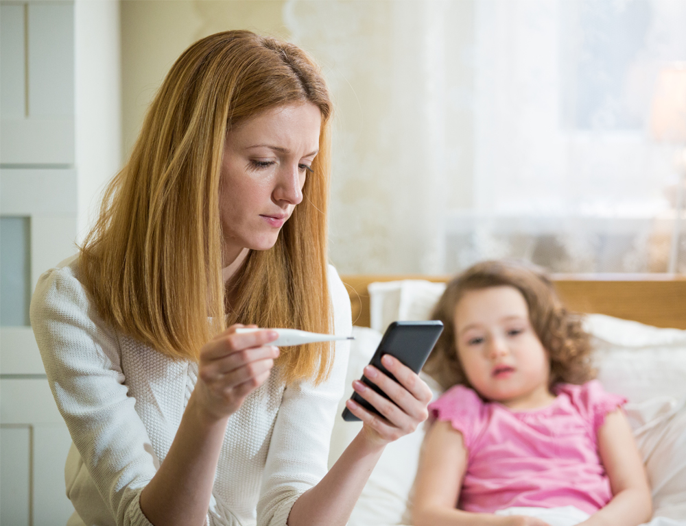 A woman holding a thermometer and a cell phone sitting next to a sick child.