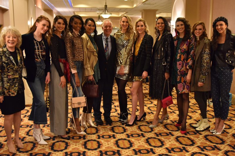 Jeanne and Ed Arnold smile with 10 models at the 2018 Neiman Marcus Runway Show. The models wear a variety of fashions and stand in a row on an ornate carpet.