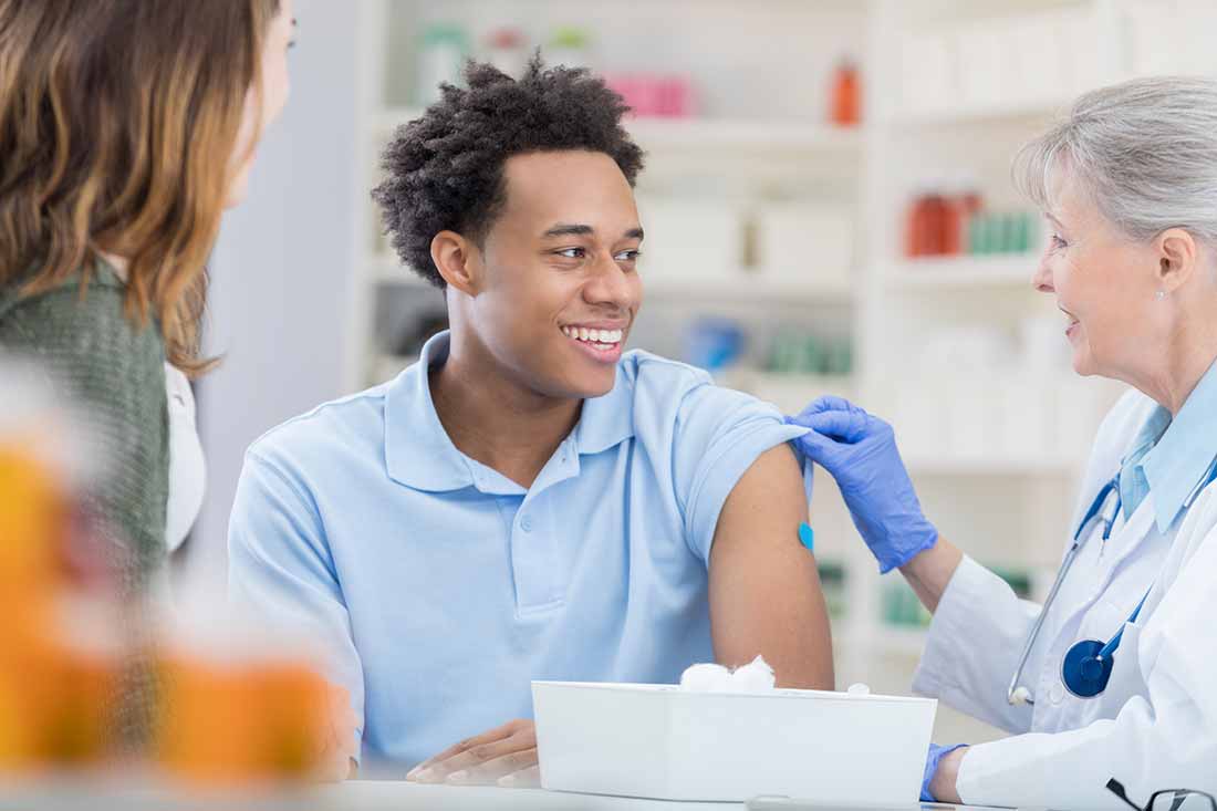 A man in the center smiles at a doctor who tugs at the sleeve of his short-sleeve shirt. A bandage on his arm. A woman on the left in the foreground looks on.