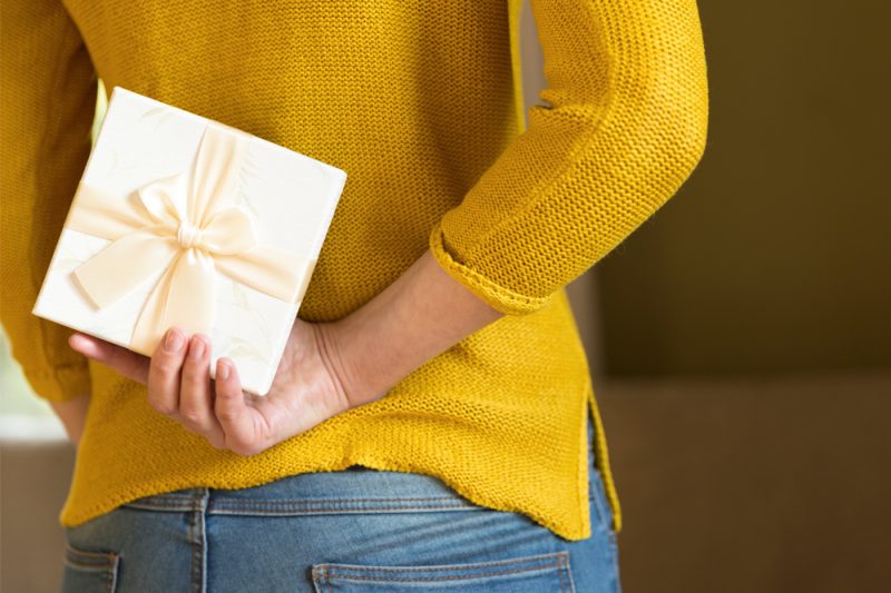A woman in a sweater and jeans is standing, holding a small, wrapped gift behind her back with her right hand.