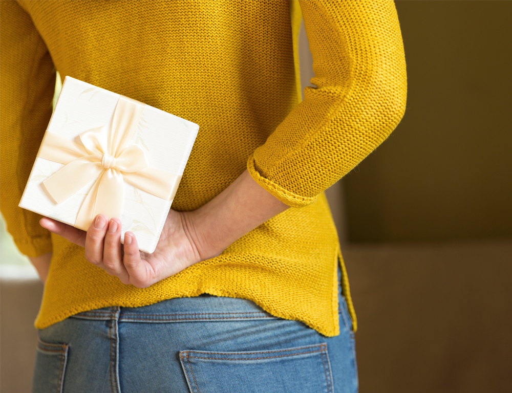 A woman in a sweater and jeans is standing, holding a small, wrapped gift behind her back with her right hand.