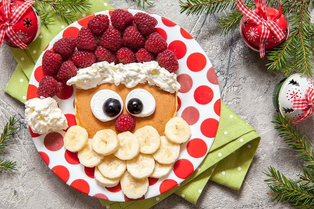 A plate surrounded by evergreens and holiday decorations features raspberries, bananas, whipped cream, blueberries and marshmallows arranged to look like the face of Santa Claus.