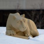 The Nittany Lion statue in the Hershey Medical Center courtyard is surrounded by snow, behind it, several trees and part of the Hershey Medical Center building are visible.