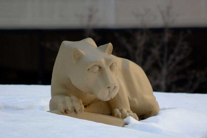 The Nittany Lion statue in the Hershey Medical Center courtyard is surrounded by snow, behind it, several trees and part of the Hershey Medical Center building are visible.