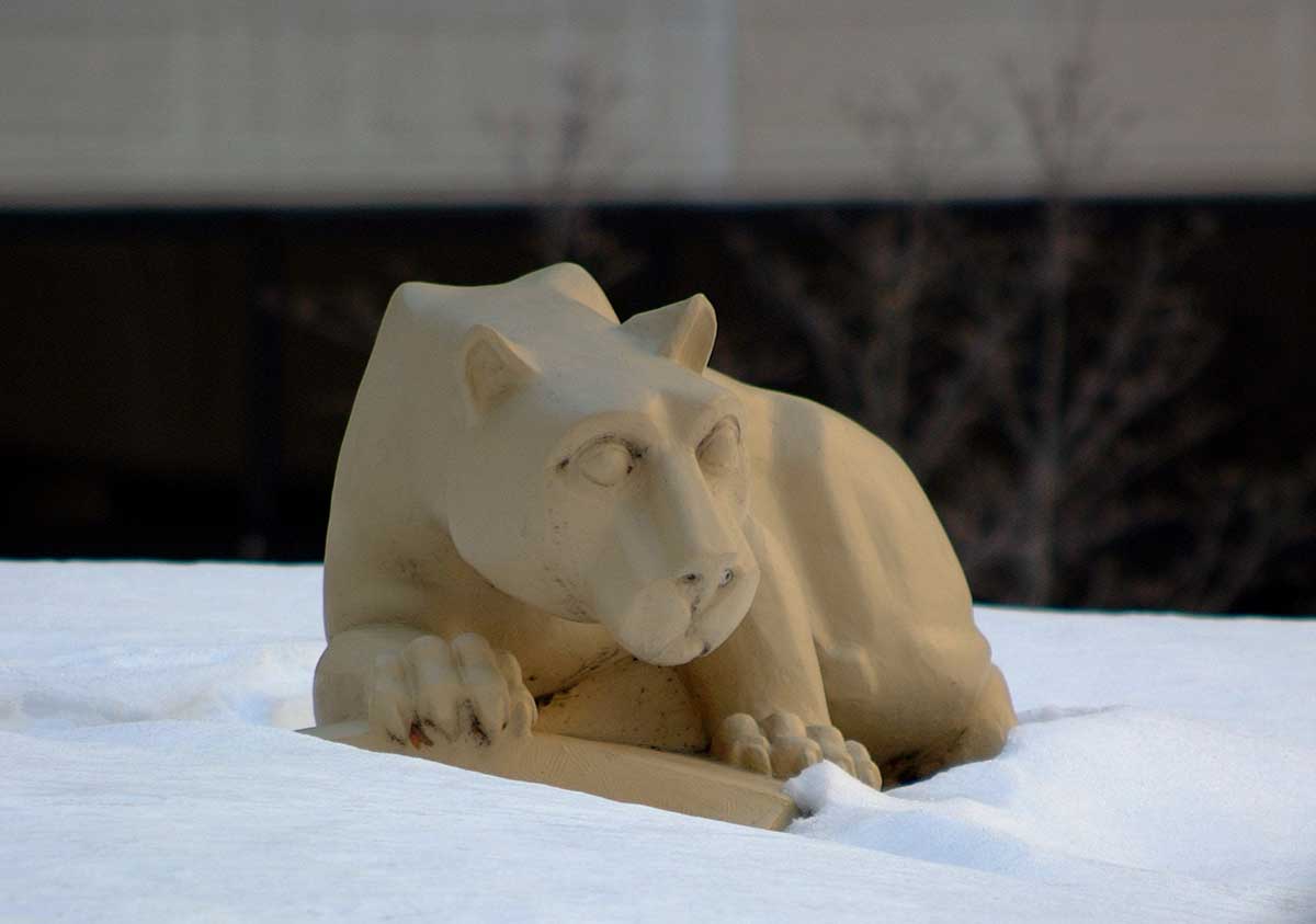 The Nittany Lion statue in the Hershey Medical Center courtyard is surrounded by snow, behind it, several trees and part of the Hershey Medical Center building are visible.