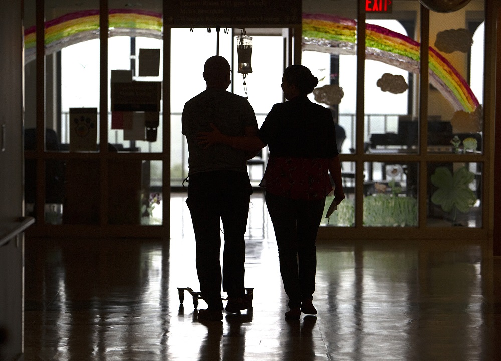 Tim Card and his wife, Tricia, walk the hallways of Penn State Cancer Institute after he receives CAR-T cell therapy infusion. Their backs are toward and the camera, and they are silhouetted against a window. Tricia puts her hand on his back. Tim is pushing an IV pole, and an IV bag with medication hangs from the pole. A rainbow is painted on the window.