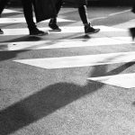 Legs and shadows cross a cross walk painted on a roadway.