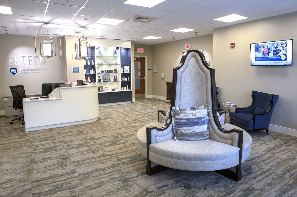 The waiting room at Esteem Penn State Health Cosmetic Associates features plush chairs, a shelf containing products behind a receptionist desk, with the Esteem logo on the wall behind it. A flat screen TV is attached to a nearby wall.