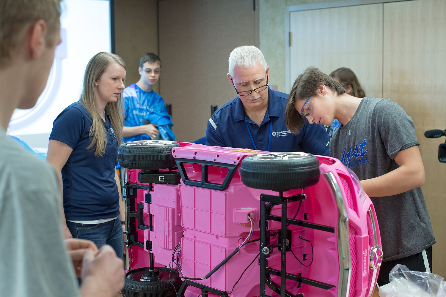 Three people work on a pink toy car, which is turned on its side and propped up on a table. Three other people stand nearby, looking on.