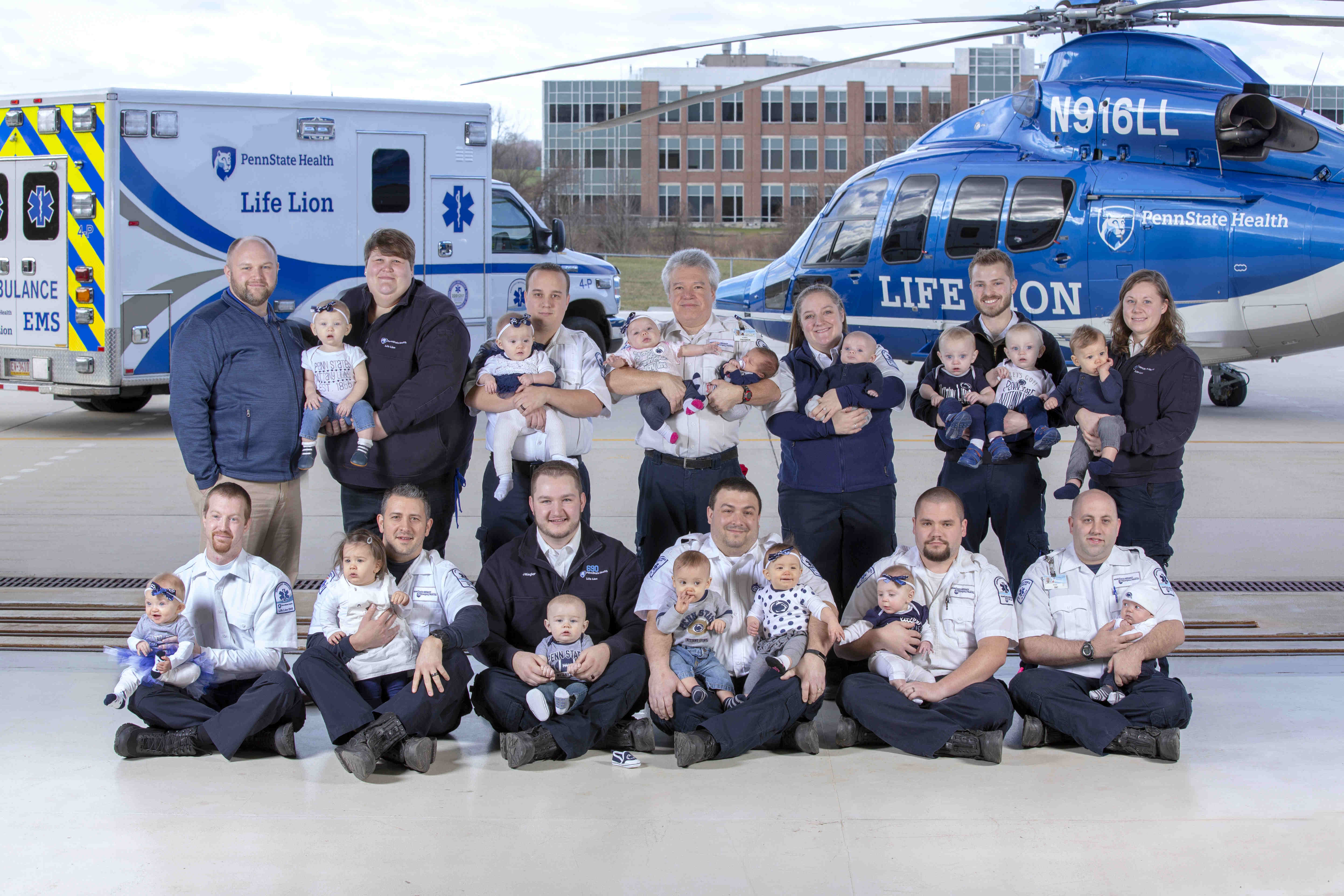 Thirteen adults pose for a photo, holding a total of 15 one-year-old children. In the near background is a Life Lion ambulance and a Life Lion helicopter. In the distant background is a brick building with dark windows.