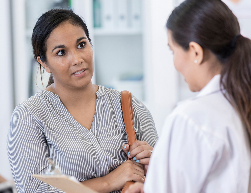 A woman in a striped shirt with a purse over her left shoulder speaks with a female doctor in a physician’s coat.