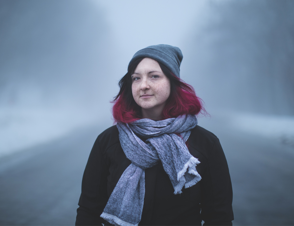 A woman in a coat, scarf and winter hat poses for a photo outdoors. She stands in the middle of a tree-lined road, which is out of focus in the background.