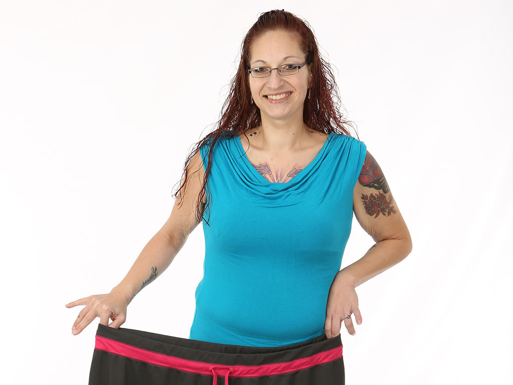Katie Rodriguez smiles as she holds the large sweatpants she used to wear before her bariatric surgery in front of her waist. She is wearing a sleeveless top and has tattoos on her arms and chest. She has long hair and glasses.
