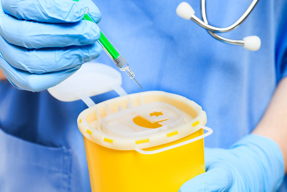A rubber-gloved hand puts a syringe in a disposal container. The person is wearing blue scrubs and a stethoscope.