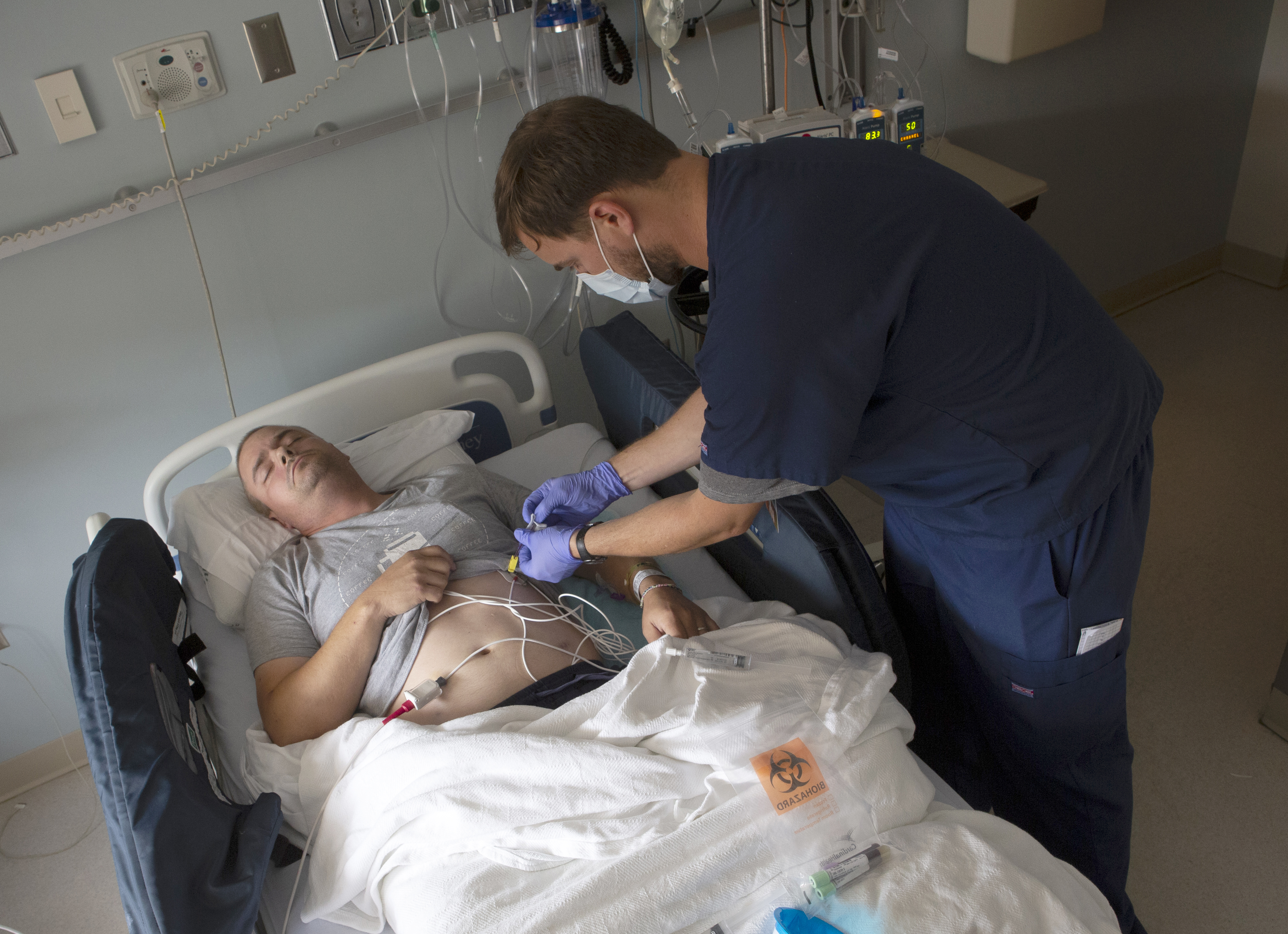 Nathan Savage-Lee, a registered nurse at Penn State Cancer Institute, leans over Tim Card and cleans his central line to prepare him for a blood draw. The line is connected to a port in his abdomen along with other lines. Tim is lying in a hospital bed with his eyes closed, holding his T-shirt up. Nathan is wearing a surgical mask and blue scrubs.